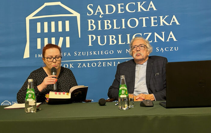 Kobieta i mężczyzna siedzą za zielonym stołem. Kobieta trzyma mikrofon i otwartą książkę. Na stole butelki z wodą oraz myszka komputerowa i laptop. Za nimi niebieski baner reklamowy z logotypem Sądeckiej Biblioteki Publicznej.