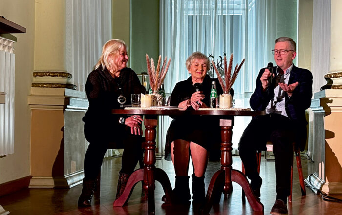 Trzy osoby ubrane na czarno siedzą na krzesłach w pomieszczeniu. Przed nimi dwa okrągłe brązowe stoliki. Na stolikach woda oraz ozdobne kwiaty. Mężczyzna po prawej stronie trzyma mikrofon. Po lewej stronie na ścianie grzejnik oraz świeca. Z tyłu okno z firanami.