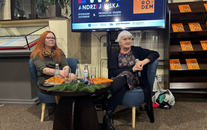 Dwie kobiety siedzą w niebieskich fotelach. Przed nimi czarny stolik z butelkami wody. Za nimi stojak z telewizorem. Na telewizorze kolorowy napis Ewa Andrzejewska. Po prawej stronie część regału z książkami.