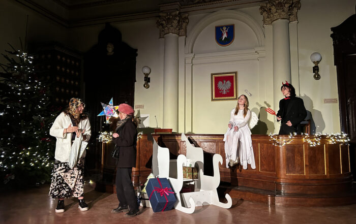 Po lewej stronie dwie osoby. Jedna ubrana na czarno, druga na biało. Na środku białe sanki i zapakowane prezenty. Po prawej stronie na brązowej ladzie siedzi dziewczyna przebrana za Anioła. Za ladą stoi chłopak przebrany za Lucyfera. Na ścianie dwie kolumny. Po bokach kinkiety oświetleniowe. Pomiędzy kolumnami wisi biały orzeł na czerwonym tle. Powyżej herb Nowego Sącza. Po lewej stronie stoi choinka z białymi lampkami.