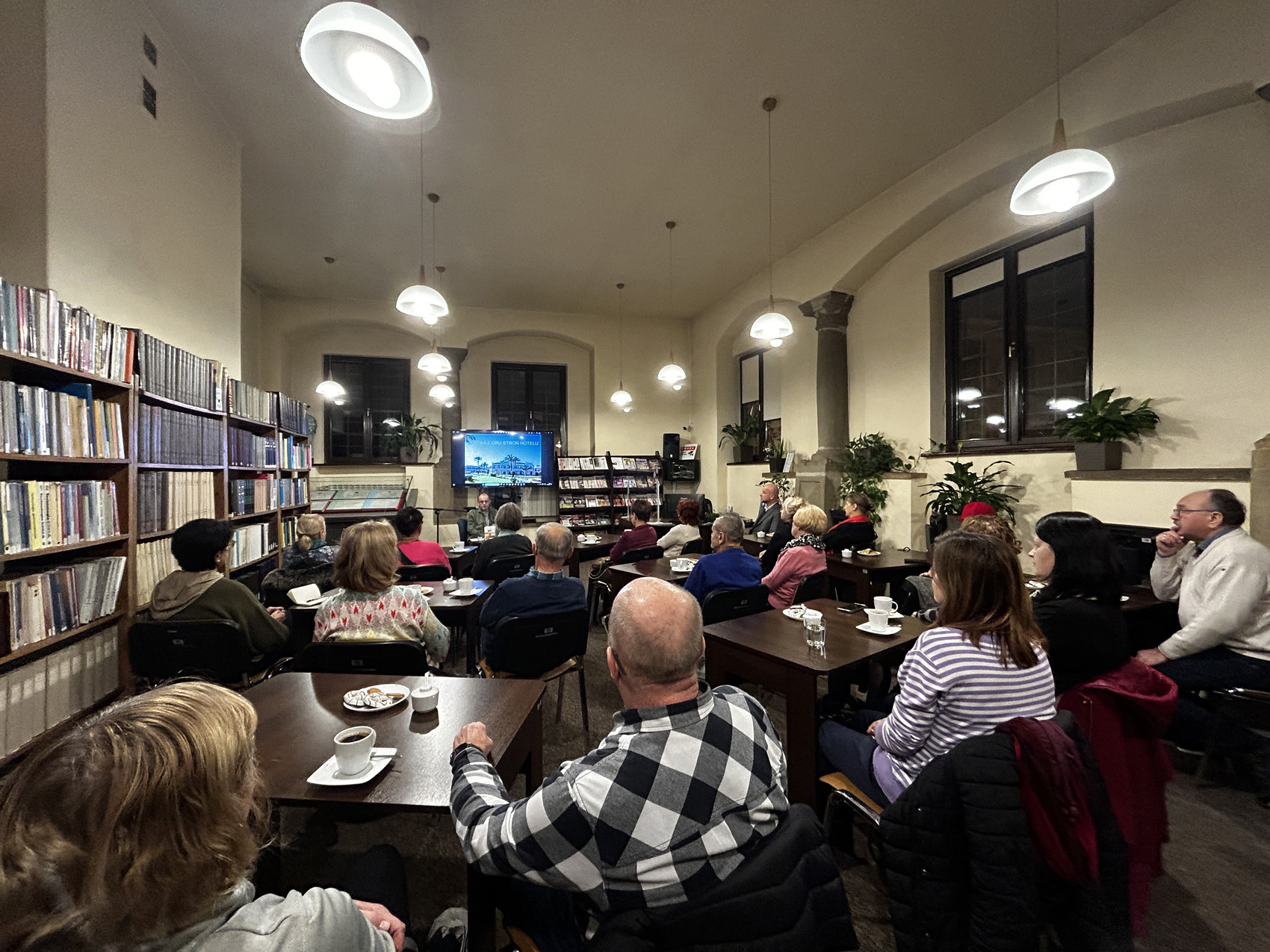 Robert Drobysz (1) Grupa ludzi siedzi na krzesłach przy brązowych stolikach w pomieszczeniu. Zaświecone są lampy sufitowe. Po lewej stronie regał z książkami. Na środku telewizor na którym wyświetlane jest zdjęcie hotelu oraz napis Afryka z obu stron hotelu. Po prawej stronie telewizora regał z czasopismami.