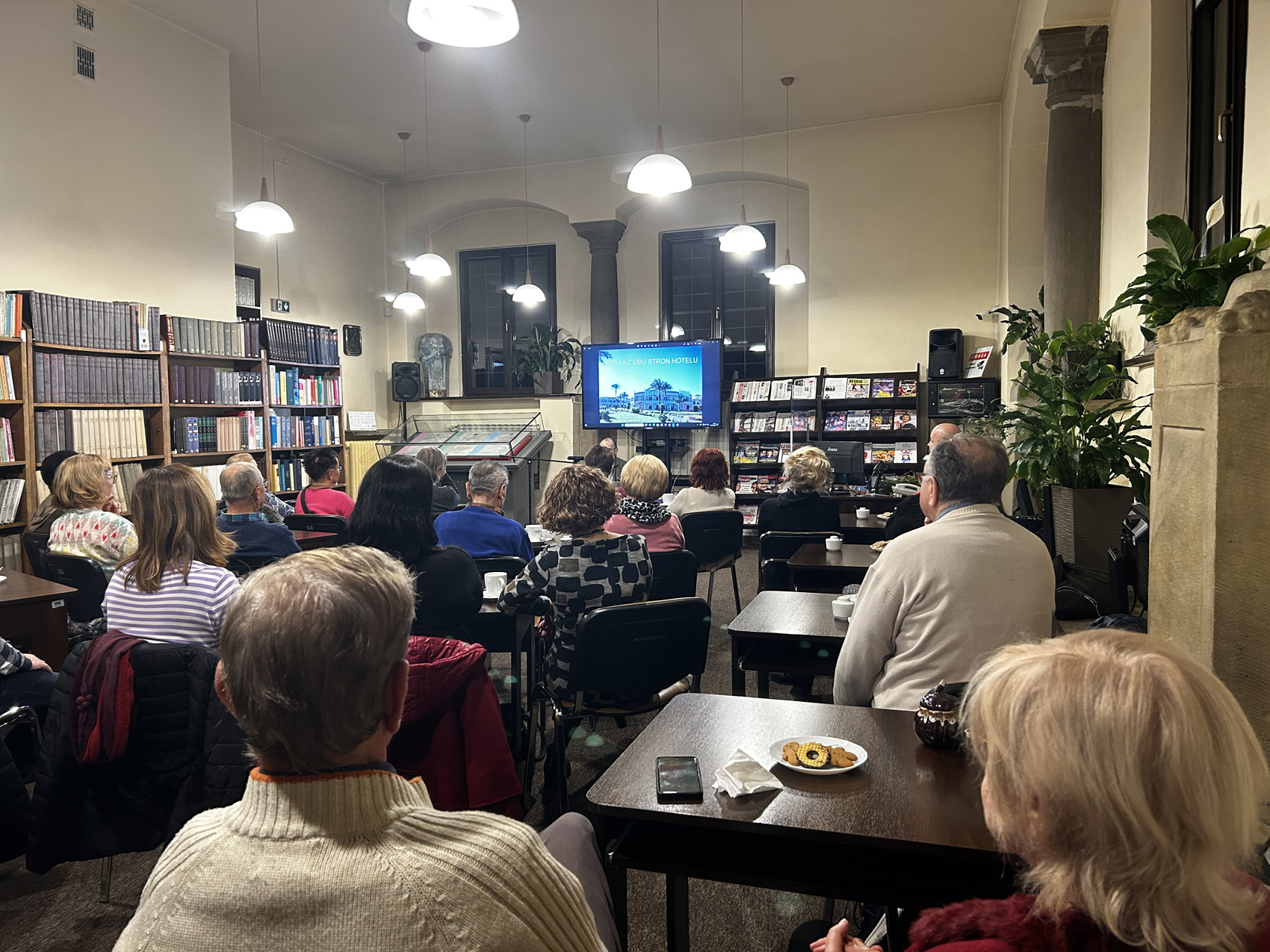 Robert Drobysz (2) Grupa ludzi siedzi na krzesłach przy brązowych stolikach w pomieszczeniu. Zaświecone są lampy sufitowe. Po lewej stronie regał z książkami. Na środku telewizor na którym wyświetlane jest zdjęcie hotelu oraz napis Afryka z obu stron hotelu. Po prawej stronie telewizora regał z czasopismami.