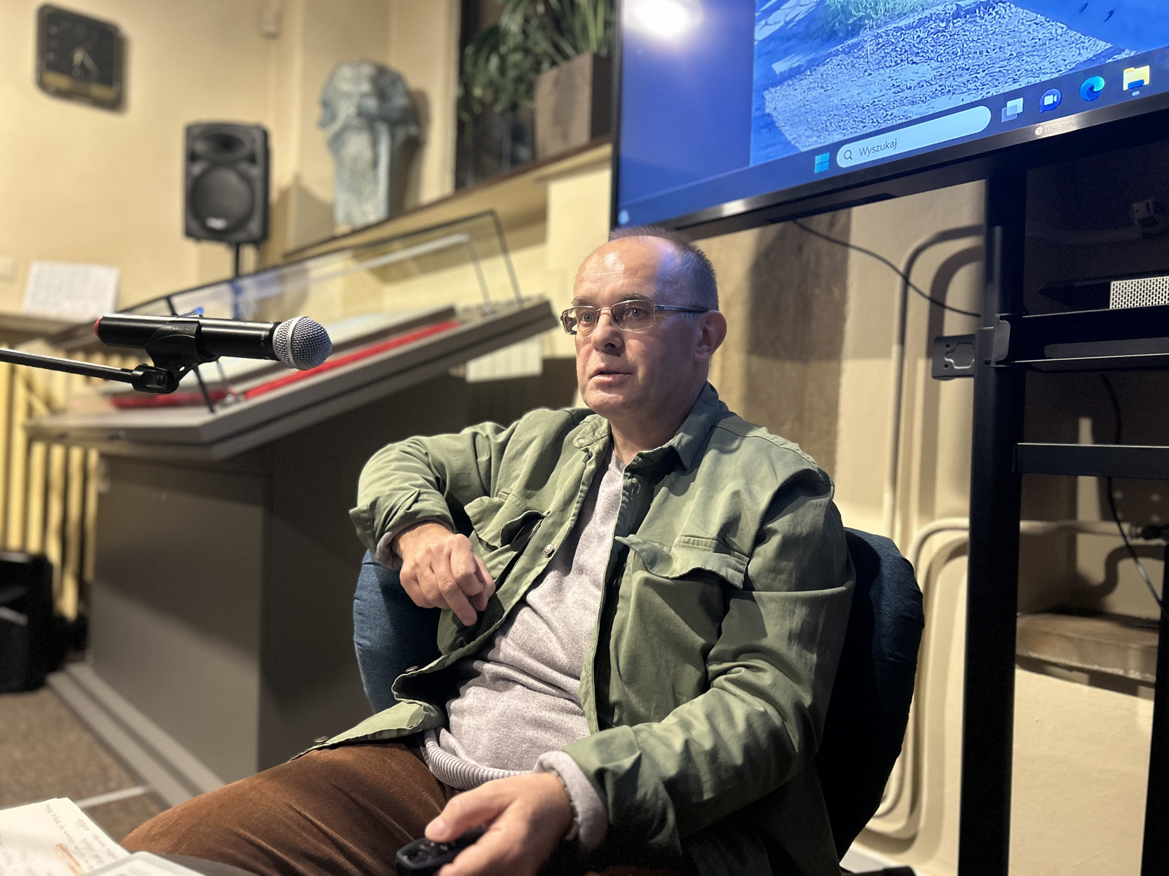 Robert Drobysz (3) Mężczyzna w zielonej kurtce siedzi na niebieskim fotelu. Po lewej stronie jest mikrofon. Na stojaku zawieszony jest telewizor. W tle szklana gablota.