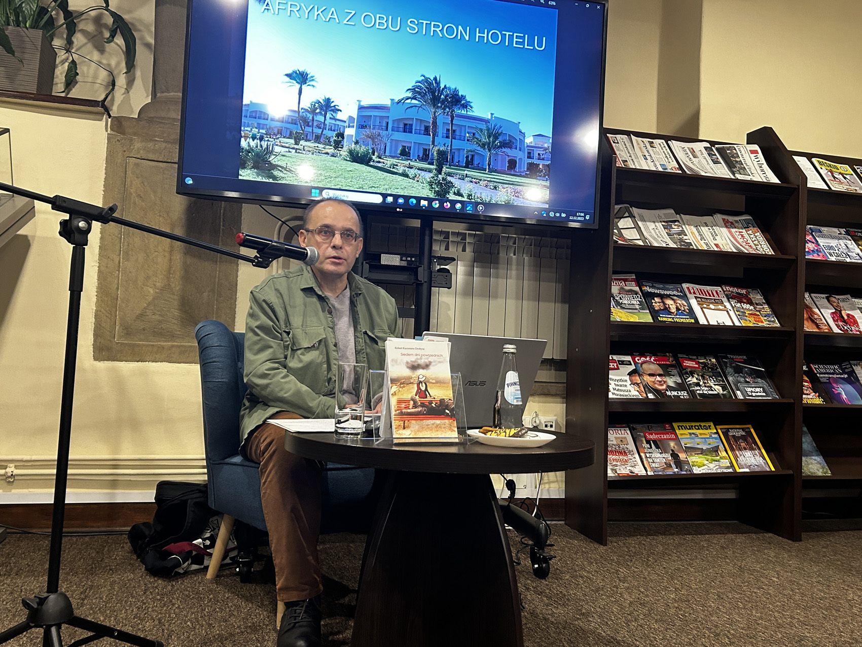 Mężczyzna w zielonej kurtce siedzi na niebieskim fotelu przy czarnym stoliku. Na stoliku jest srebrny laptop, butelka wody oraz książka. Obok statyw z mikrofonem. Za nim na stojaku telewizor. Na telewizorze wyświetlane jest zdjęcie hotelu oraz napis Afryka z obu stron hotelu. Po prawej stronie regał z czasopismami.