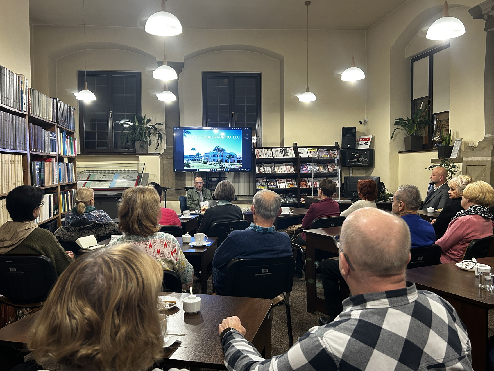 Robert Drobysz (6) Grupa ludzi siedzi na krzesłach przy brązowych stolikach w pomieszczeniu. Zaświecone są lampy sufitowe. Po lewej stronie regał z książkami. Na środku telewizor na którym wyświetlane jest zdjęcie hotelu oraz napis Afryka z obu stron hotelu. Po prawej stronie telewizora regał z czasopismami.