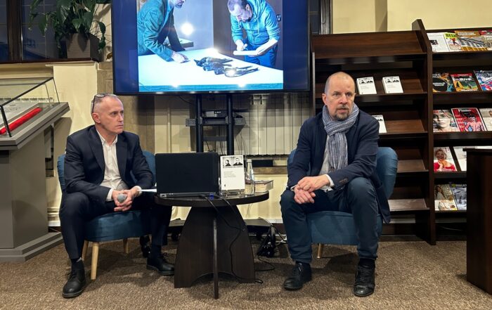 W pomieszczeniu znajduje się dwóch mężczyzn. Mężczyźni siedzą na fotelach. Pomiędzy nimi stoi mały stolik. Na stoliku znajdują się: laptop, książka, butelka z wodą. Mężczyzna po lewej stronie trzyma w rękach mikrofon. Za mężczyznami stoi duży ekran. Na ekranie wyświetla się zdjęcie. Po lewej stronie przy ścianie stoi długi brązowy regał z książkami i czasopismami.