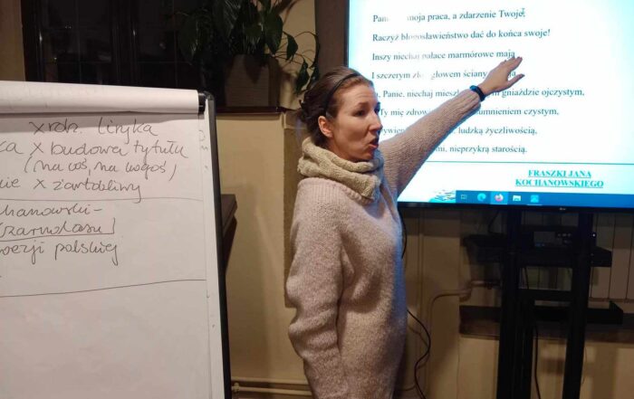 Kobieta w kremowym długim swetrze wskazuje ręką na tekst wyświetlony na dużym ekranie. Obok flipchart z zapisanymi czarnym flamastrem arkuszami papieru.