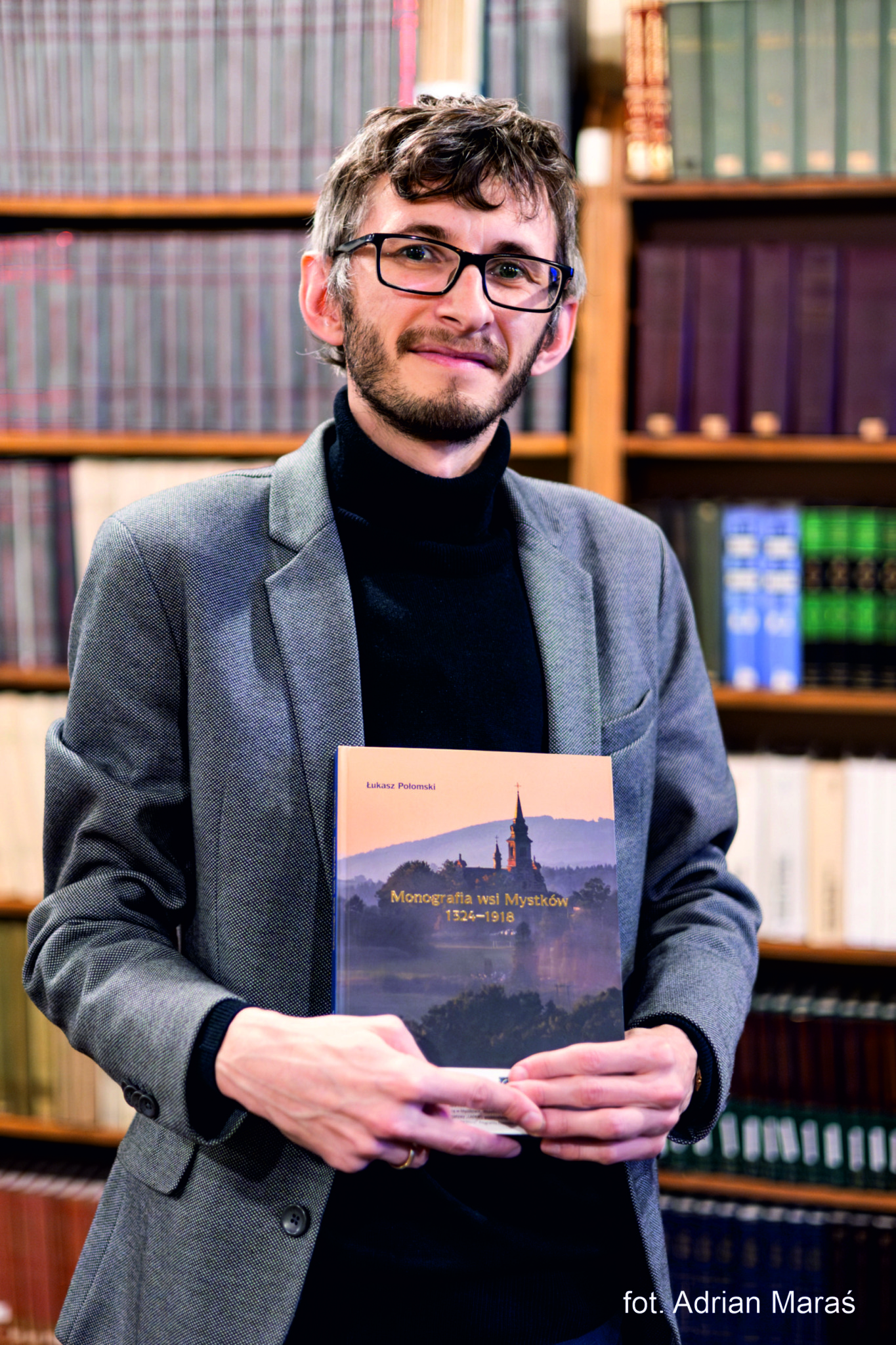 Łukasz Połomski Foto Mężczyzna w okularach, w golfie i szarej marynarce lekko się uśmiecha. W dłoniach trzyma książkę. Na okładce książki zdjęcie kościoła i napis "Monografia wsi Mystków 1324-1918". Za mężczyzną regały z książkami.