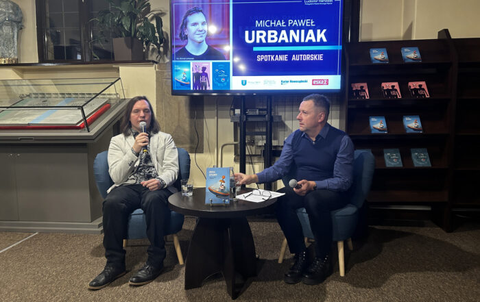 Dwóch mężczyzn siedzi przy małym brązowym stoliku. Trzymają w rękach mikrofony. Za nimi po lewej stronie gablota z wyeksponowanymi księgami, po prawej stronie regał z książkami. W tle duży telewizor z kolorową grafiką.