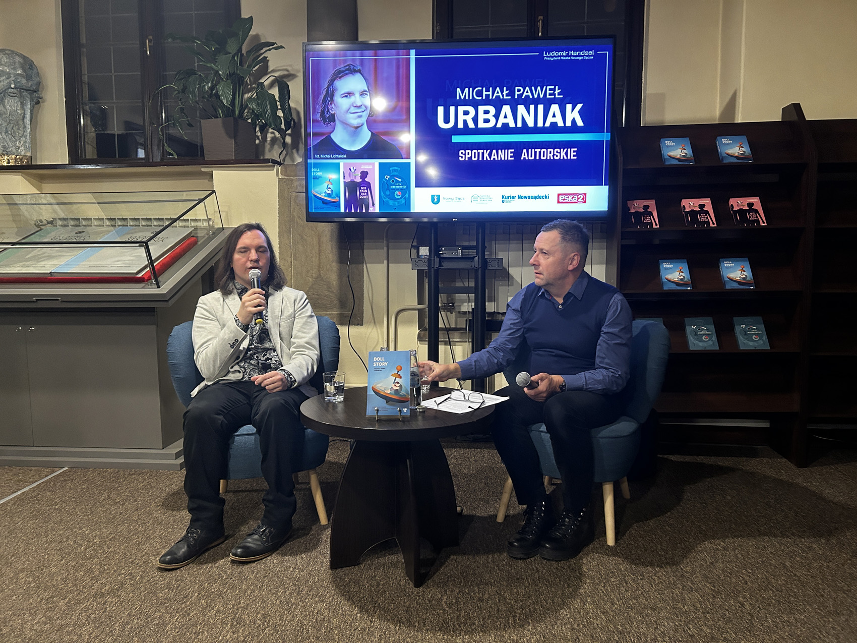 Dwóch mężczyzn siedzi przy małym brązowym stoliku. Trzymają w rękach mikrofony. Za nimi po lewej stronie gablota z wyeksponowanymi księgami, po prawej stronie regał z książkami. W tle duży telewizor z kolorową grafiką.