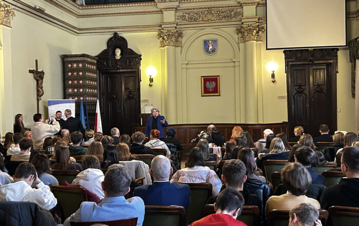 Sala Ratuszowa. Na krzesłach siedzi grupa około 50 osób, pokazanych od tyłu. Przed nimi na środku stoi starsza kobieta. Przed kobietą stoi na statywie mikrofon. Kobieta mówi do mikrofonu. Po lewej stronie pod ścianą stoi grupa 4 osób. Po prawej stronie w górnym rogu wisi ekran.