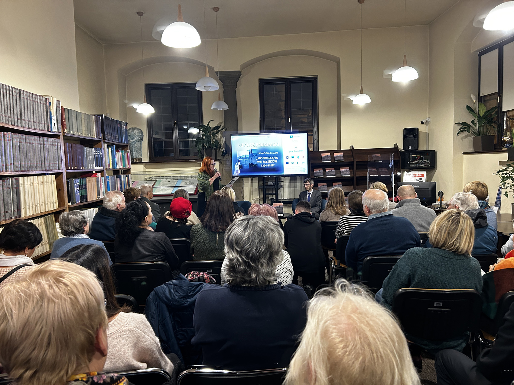 Promocja ksiażki Łukasza Połomskiego (1) W pomieszczeniu znajduje się grupa 27 siedzących osób, pokazanych od tyłu. Przed siedzącymi na krzesłach ludźmi na środku stoi kobieta. Kobieta trzyma w jednej ręce kartkę, w drugiej mikrofon. Czyta. Obok kobiety na fotelu siedzi młody mężczyzna. W tle duży ekran. Na ekranie wyświetlone zdjęcie. Po lewej stronie obok siedzących stoi długi regał z książkami.