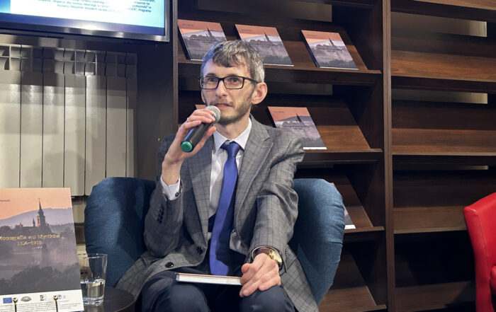 W pomieszczeniu znajduje się mężczyzna siedzący na granatowym fotelu. Mężczyzna ubrany jest w szarą marynarkę w kratkę. W prawej ręce trzyma mikrofon blisko ust, mówi do niego. Obok mężczyzny po lewej stronie stoi okrągły stolik a na nim książka i szklanka z wodą. Po prawej stronie za mężczyzną stoi duży brązowy regał. Na regale kilka książek.