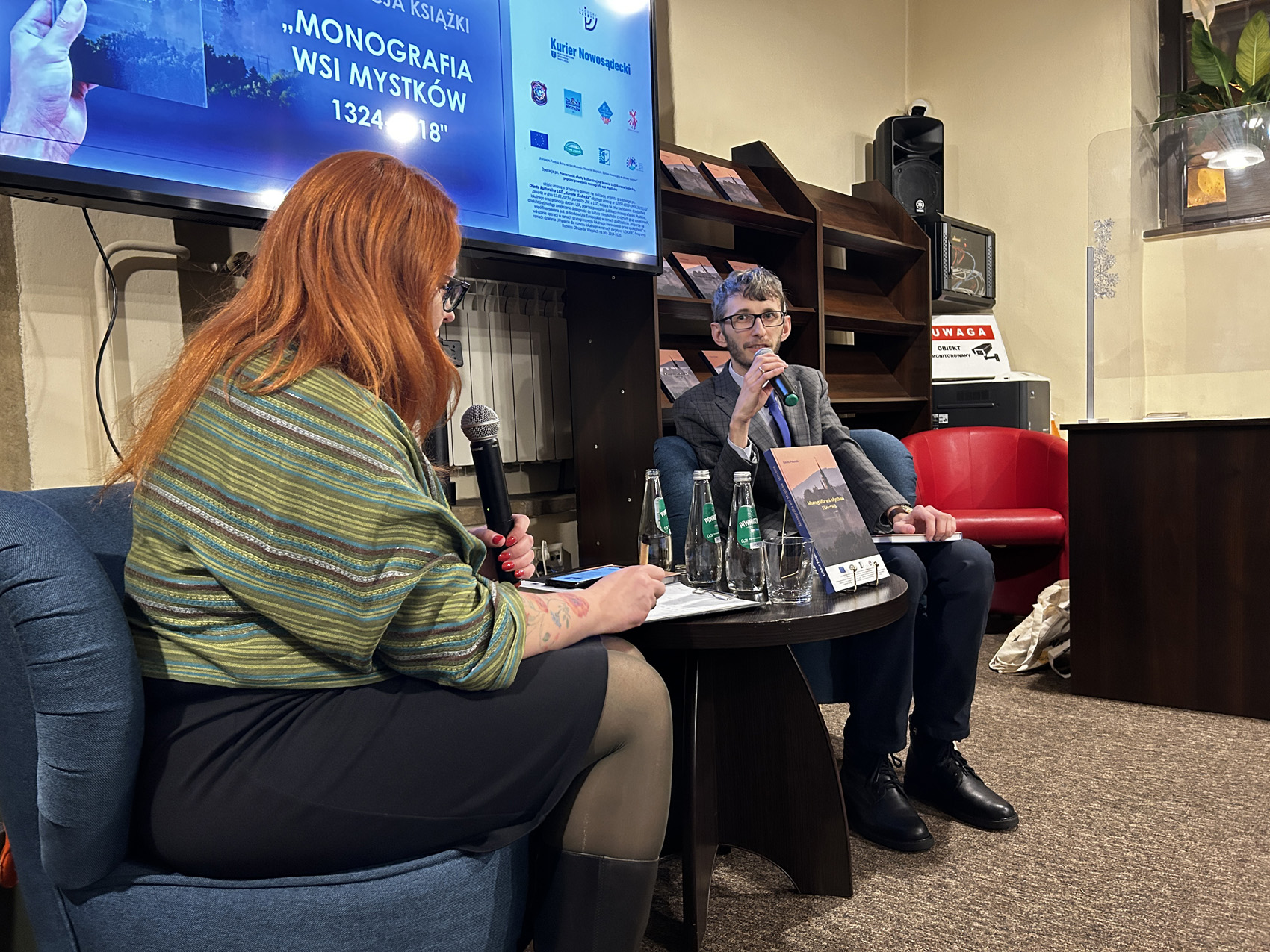 Promocja ksiażki Łukasza Połomskiego (3) W pomieszczeniu znajdują się dwie osoby: kobieta i mężczyzna. Po lewej stronie na fotelu siedzi kobieta pokazana bokiem. Ubrana jest w czarną spódnicę do kolana. Na ramionach ma zielono-żółty szal. W lewej ręce trzyma mikrofon. Obok niej stoi mały okrągły stolik. Na stoliku są: książka, kartki papieru, szklanka, trzy butelki wody. Za stolikiem na fotelu po prawej stronie siedzi młody mężczyzna. Mężczyzna trzyma w ręce mikrofon blisko ust, mówi do niego. Za siedzącymi stoi duży ekran. Na ekranie wyświetla się zdjęcie. Obok ekranu po prawej stronie brązowy regał, a na nim książki.