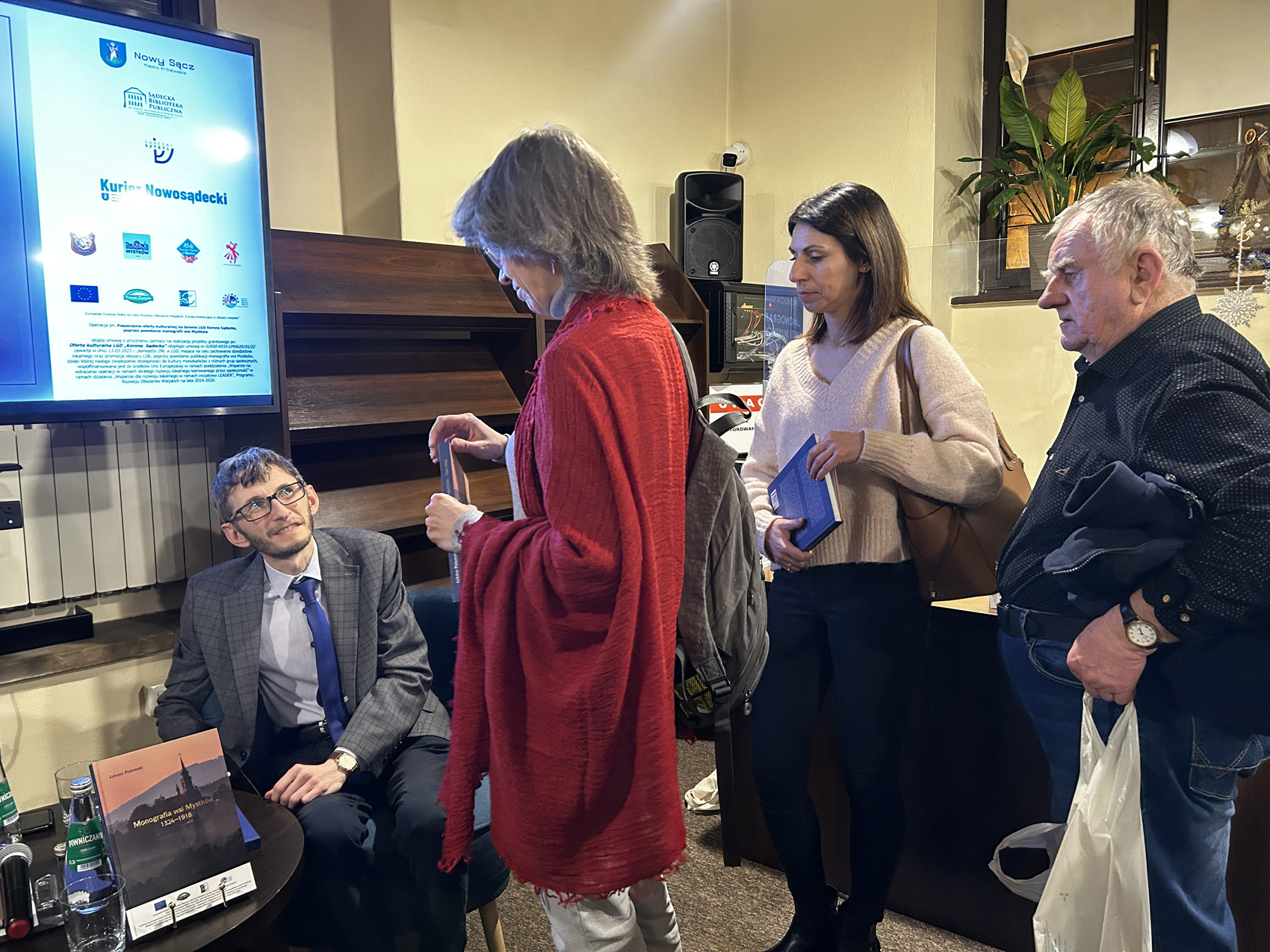 Promocja ksiażki Łukasza Połomskiego (4) W pomieszczeniu znajdują się cztery osoby: dwóch mężczyzn i dwie kobiety. Po lewej stronie na fotelu przy okrągłym stoliku siedzi mężczyzna w szarej marynarce. Przed mężczyzną stoją trzy osoby: dwie kobiety i mężczyzna. Kobieta pierwsza od lewej ma na sobie czerwoną narzutkę. Pokazana jest z boku. W rękach trzyma książkę. Siedzący mężczyzna patrzy na nią. Za kobietą stoi druga kobieta ubrana w ciemne spodnie i kremowy sweterek. Kobieta także trzyma w rękach książkę. Za kobietą po prawej stronie stoi mężczyzna pokazany z boku. Mężczyzna trzyma w prawej ręce reklamówkę.