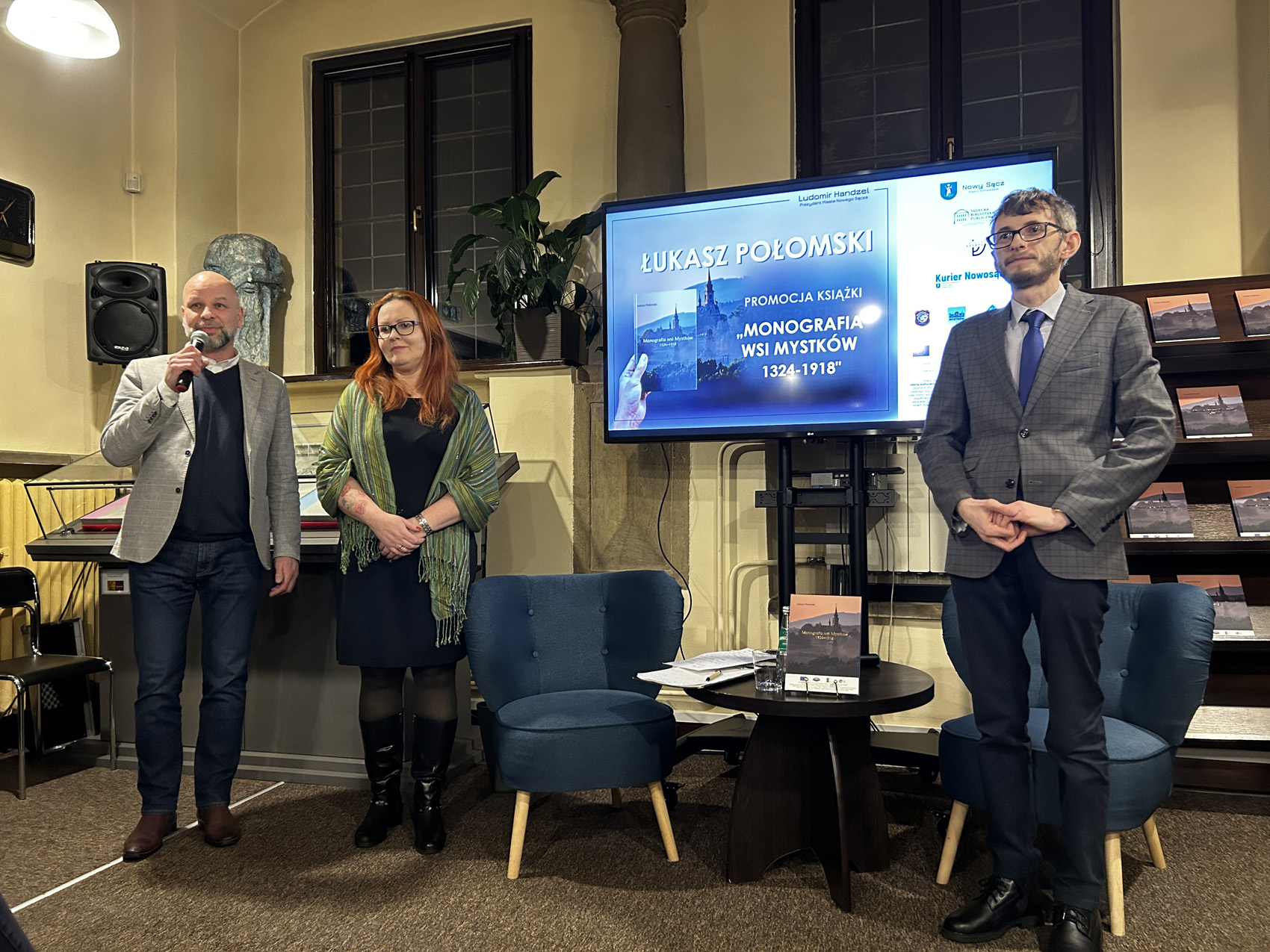 Promocja ksiażki Łukasza Połomskiego (5) W pomieszczeniu znajdują się trzy osoby: dwóch mężczyzn i kobieta. Po lewej stronie stoi mężczyzna w szarej marynarce. Mężczyzna trzyma w jednej ręce mikrofon blisko ust, mówi do niego. Obok mężczyzny stoi kobieta. Obok kobiety stoją dwa granatowe fotele, a między nimi mały okrągły stolik. Na stoliku są: książka, kartki papieru, szklanka. Przed fotelem po prawej stronie stoi młody mężczyzna w ciemnoszarej marynarce. Za nimi w tle duży ekran. Na ekranie wyświetla się zdjęcie. Obok ekranu po prawej stronie brązowy regał, a na nim książki.