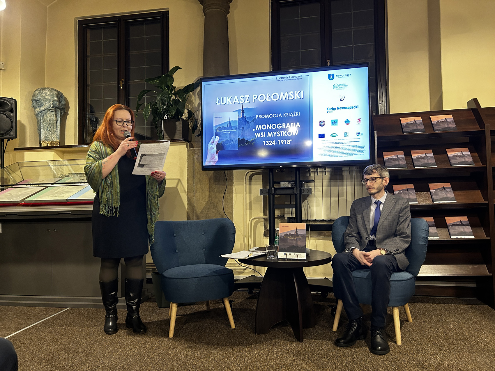 Promocja ksiażki Łukasza Połomskiego (6) W pomieszczeniu znajdują się dwie osoby: kobieta i mężczyzna. Kobieta stoi po lewej stronie. W jednej ręce trzyma kartkę, w drugiej mikrofon blisko ust. Kobieta patrzy na kartkę, czyta. Obok kobiety stoją dwa granatowe fotele, a między nimi mały okrągły stolik. Na stoliku są: książka, kartki papieru, szklanka. Na fotelu po prawej stronie siedzi młody mężczyzna. Za nimi duży ekran. Na ekranie wyświetla się zdjęcie. Obok ekranu po prawej stronie brązowy regał, a na nim książki.