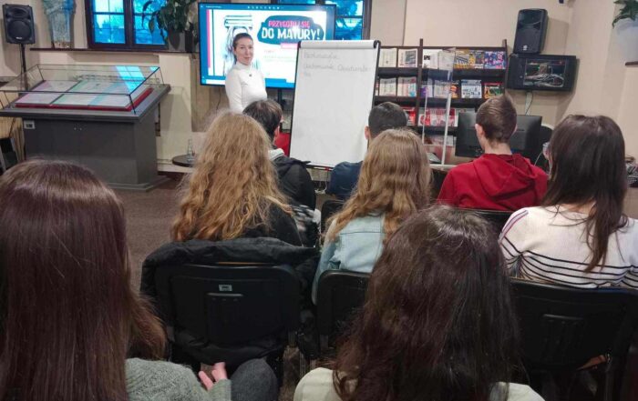 Wypełniona sala. Widownia siedzi na krzesłach. Przed nią uśmiechnięta kobieta w białej bluzce. Stoi przy flipcharcie. W tle duży telewizor z kolorową grafiką.