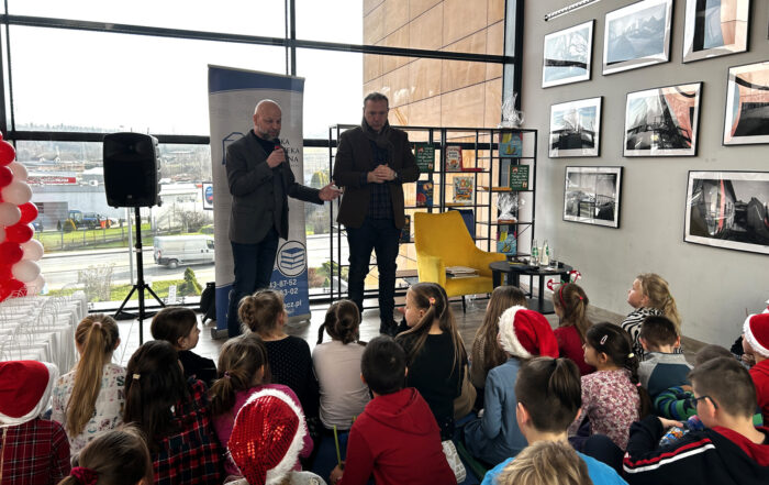 Na pierwszym planie grupa dzieci tyłem do fotografującego. Przed nimi centralnie dwóch mężczyzn. Jeden trzyma mikrofon. Po prawej stronie na ścianie zdjęcia.