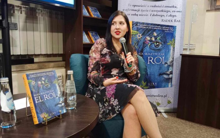 Kobieta w kwiecistej sukience siedzi na niebieskim fotelu, mówiąc do mikrofonu. Po prawej stronie ustawiony jest baner z wizerunkiem książki ora napisem Beata Skrzypczak El Roi. Po lewej stronie znajduje się brązowy stolik z butelkami wody oraz książką w niebieskiej okładce: Beata Skrzypczak, El Roi.