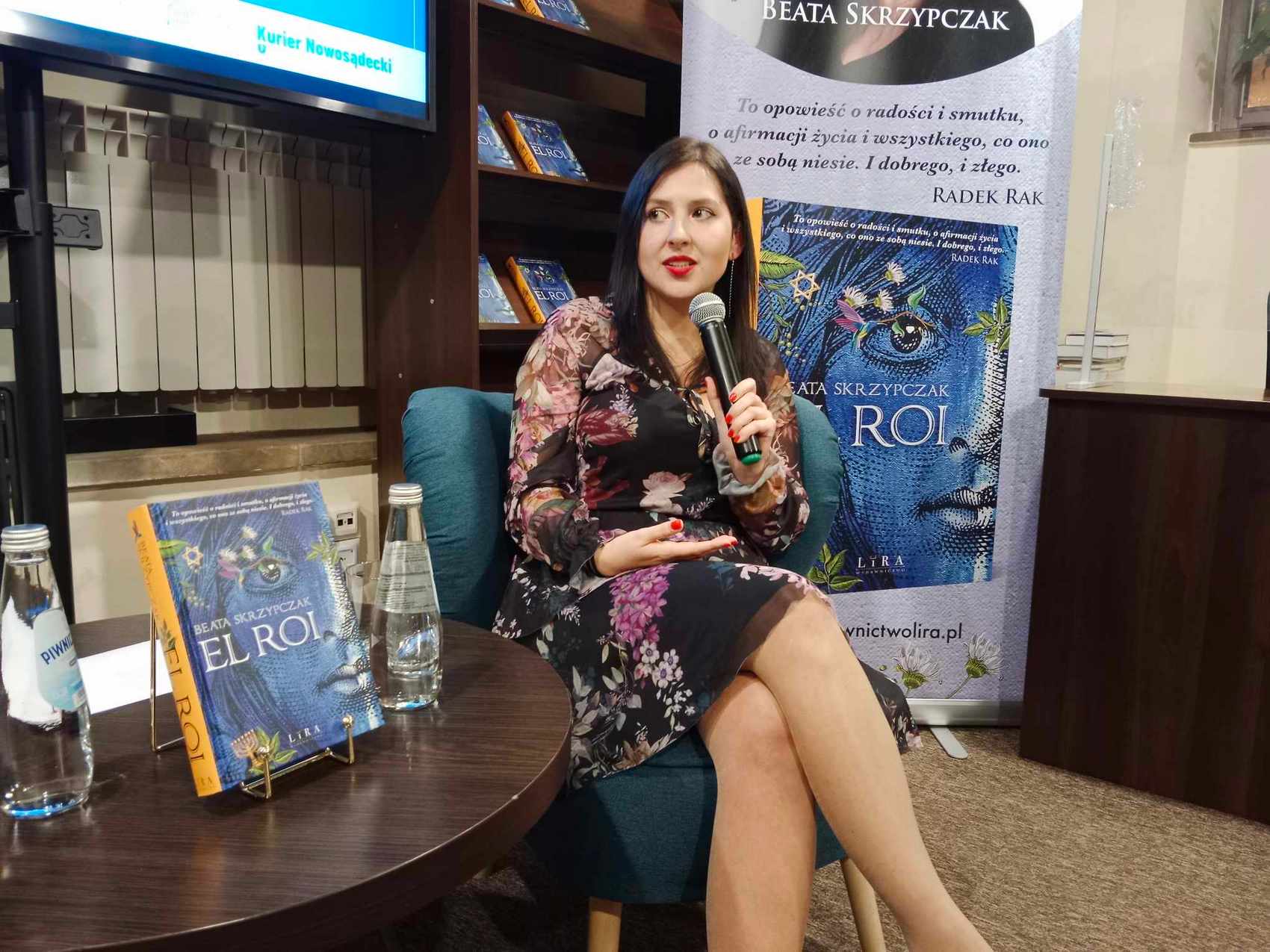 Kobieta w kwiecistej sukience siedzi na niebieskim fotelu, mówiąc do mikrofonu. Po prawej stronie ustawiony jest baner z wizerunkiem książki ora napisem Beata Skrzypczak El Roi. Po lewej stronie znajduje się brązowy stolik z butelkami wody oraz książką w niebieskiej okładce: Beata Skrzypczak, El Roi.