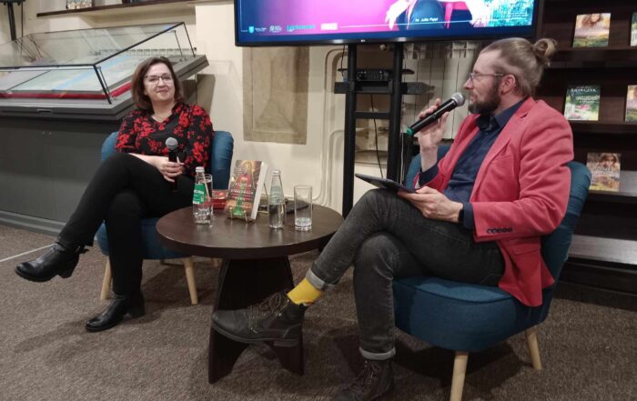 Kobieta i mężczyzna siedzą przy okrągłym, brązowym stoliku, na którym stoją napoje oraz książka. Kobieta trzyma mikrofon a mężczyzn mówi do mikrofonu.