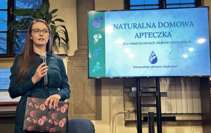 Kobieta prowadzi wykład z mikrofonem w ręce, obok ustawiony jest ekran, na którym widnieją właściwości olejków eterycznych.