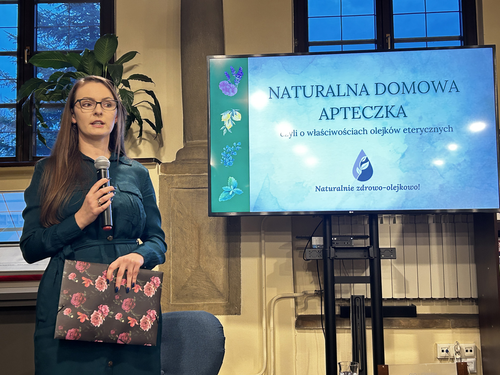 Kobieta prowadzi wykład z mikrofonem w ręce, obok ustawiony jest ekran, na którym widnieją właściwości olejków eterycznych.