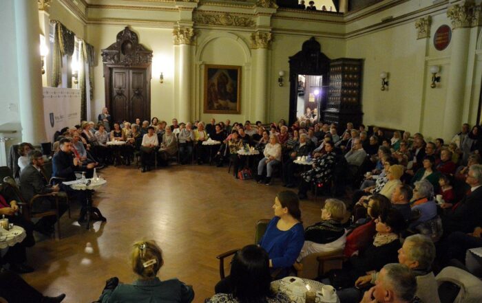 Na zdjęciu widać licznie zgromadzonych uczestników spotkania w eleganckiej sali Ratusza, Wszyscy spoglądają na przemawiającego mężczyznę siedzącego przy stoliku.