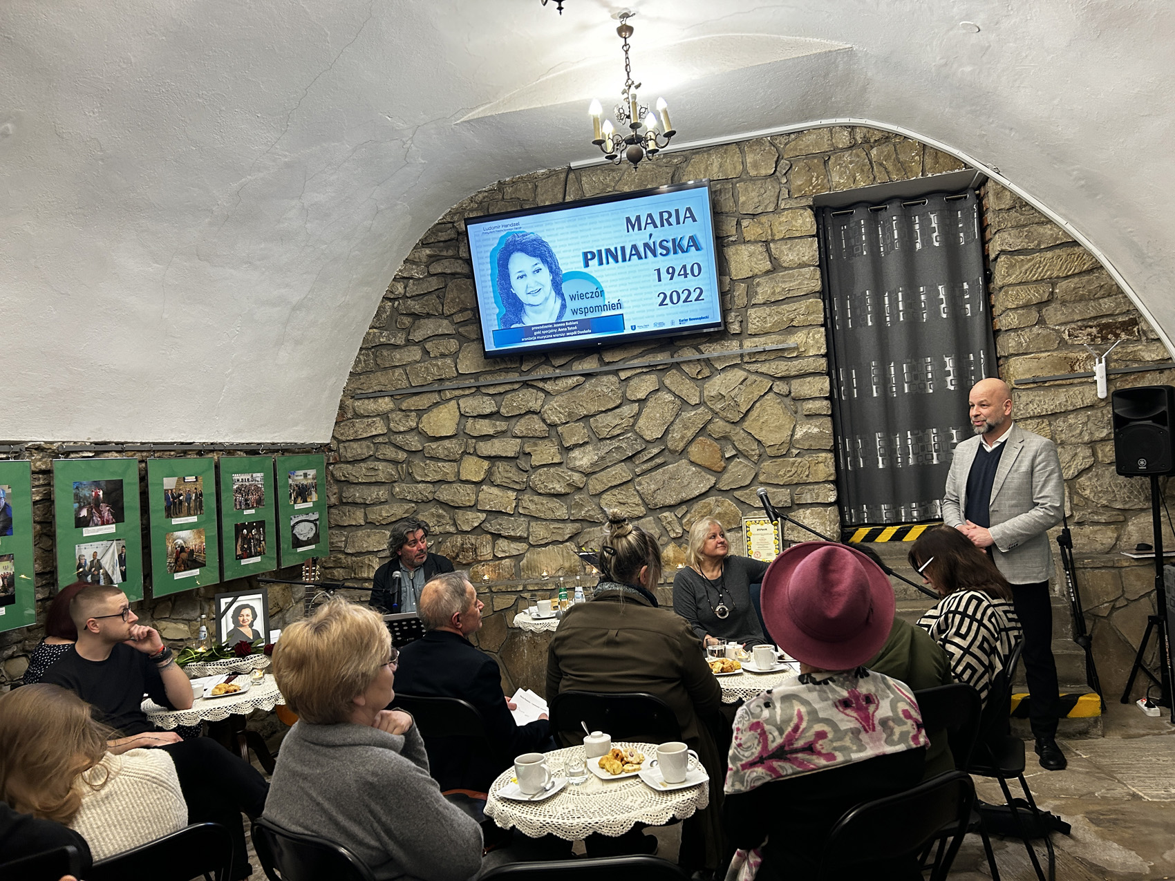 Wieczór wspomnień o Marii Piniańskiej (1) Dyrektor biblioteki przemawia, zgromadzeni słuchają go siedząc przy stolikach. Na kamiennych ścianach rozwieszone są zdjęcia oraz ekran z wizerunkiem Marii Piniańskiej oraz data jej urodzin i śmierci 1940 - 2022.