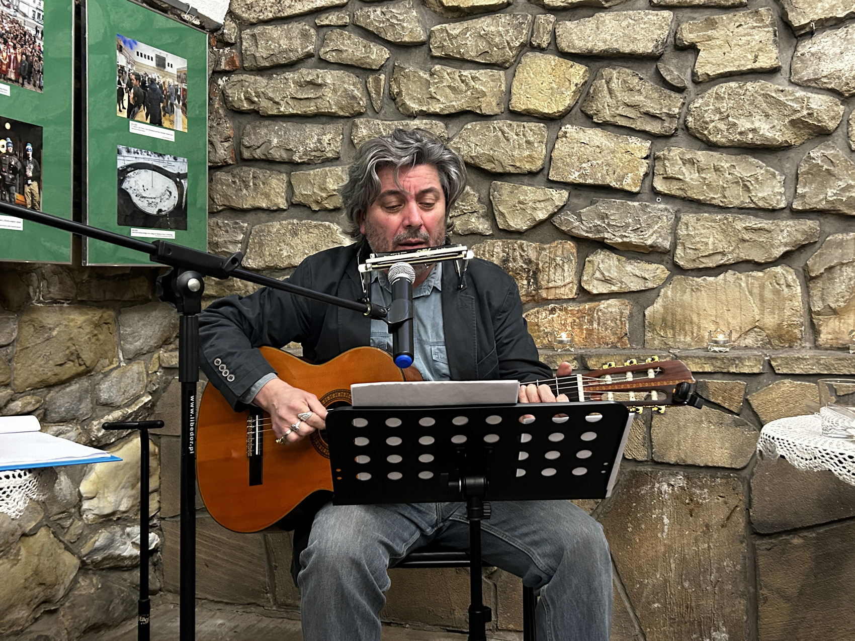 Wieczór wspomnień o Marii Piniańskiej (2) Mężczyzna śpiewa i gra na gitarze patrząc na tekst piosenki. W tle kamienna ściana oraz zdjęcia.