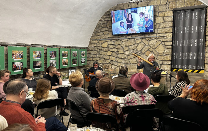 Zebrani słuchają mężczyzny, śpiewającego i grającego na gitarze. Za nim na ekranie wyświetlają się zdjęcia Marii Piniańskiej.