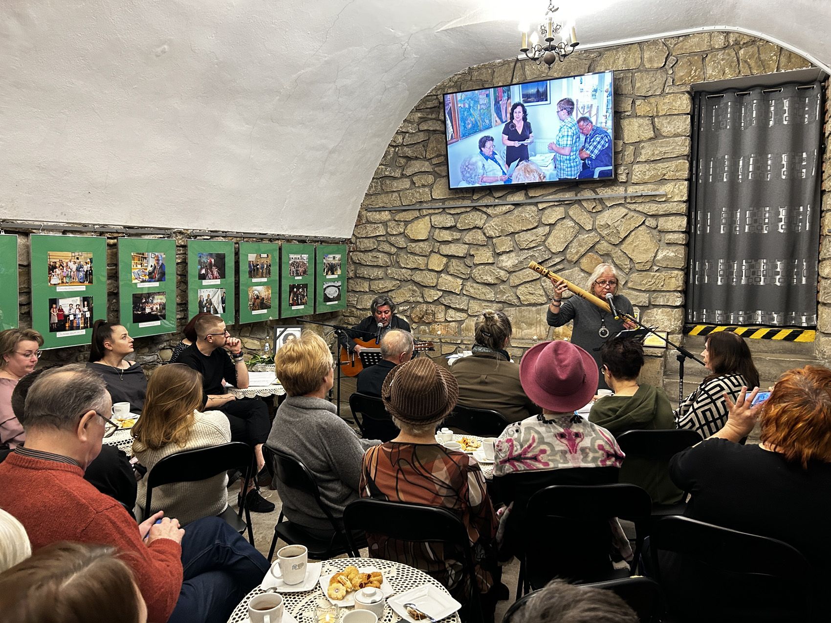 Zebrani słuchają mężczyzny, śpiewającego i grającego na gitarze. Za nim na ekranie wyświetlają się zdjęcia Marii Piniańskiej.