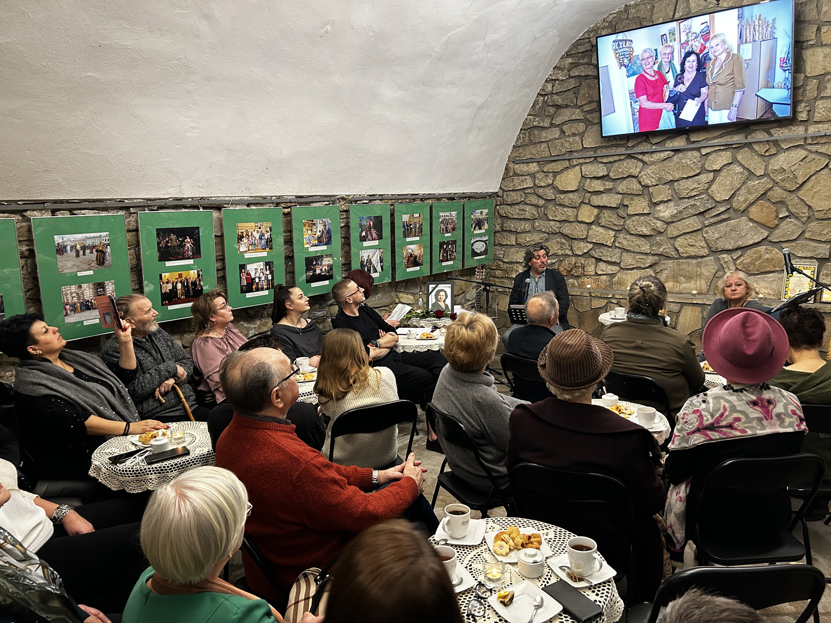 Wieczór wspomnień o Marii Piniańskiej (5) Uczestnicy spotkania oglądają zdjęcia Marii Piniańskiej, na kamiennych ścianach wiszą zdjęcia.
