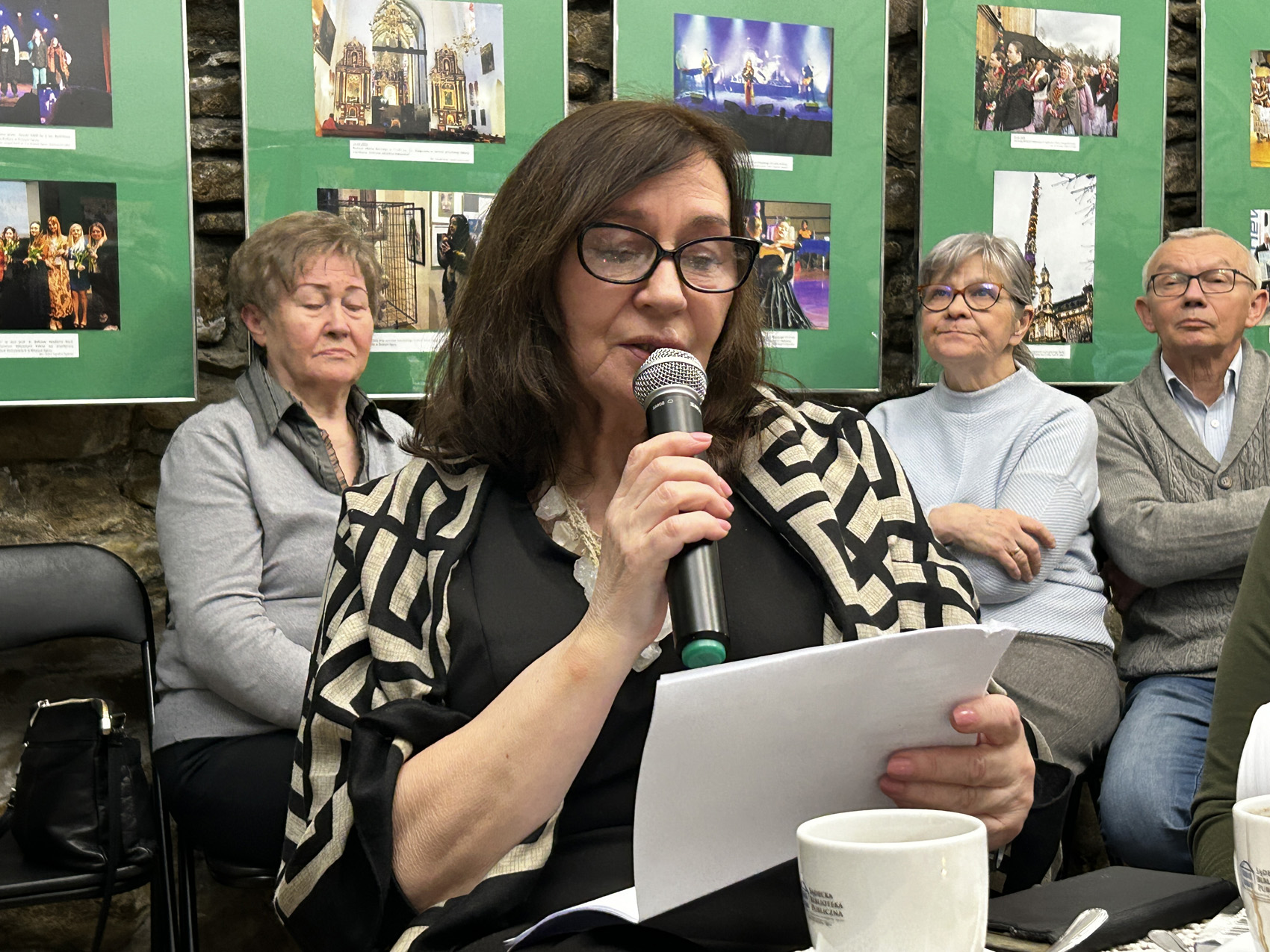 Wieczór wspomnień o Marii Piniańskiej (8) Uczestniczka spotkania czyta z kartki do mikrofonu, przed nią stoi filiżanka na stoliku, za nią widoczna jest trójka uczestników spotkania. Na ścianach wiszą zdjęcia związane z Marią Piniańską.