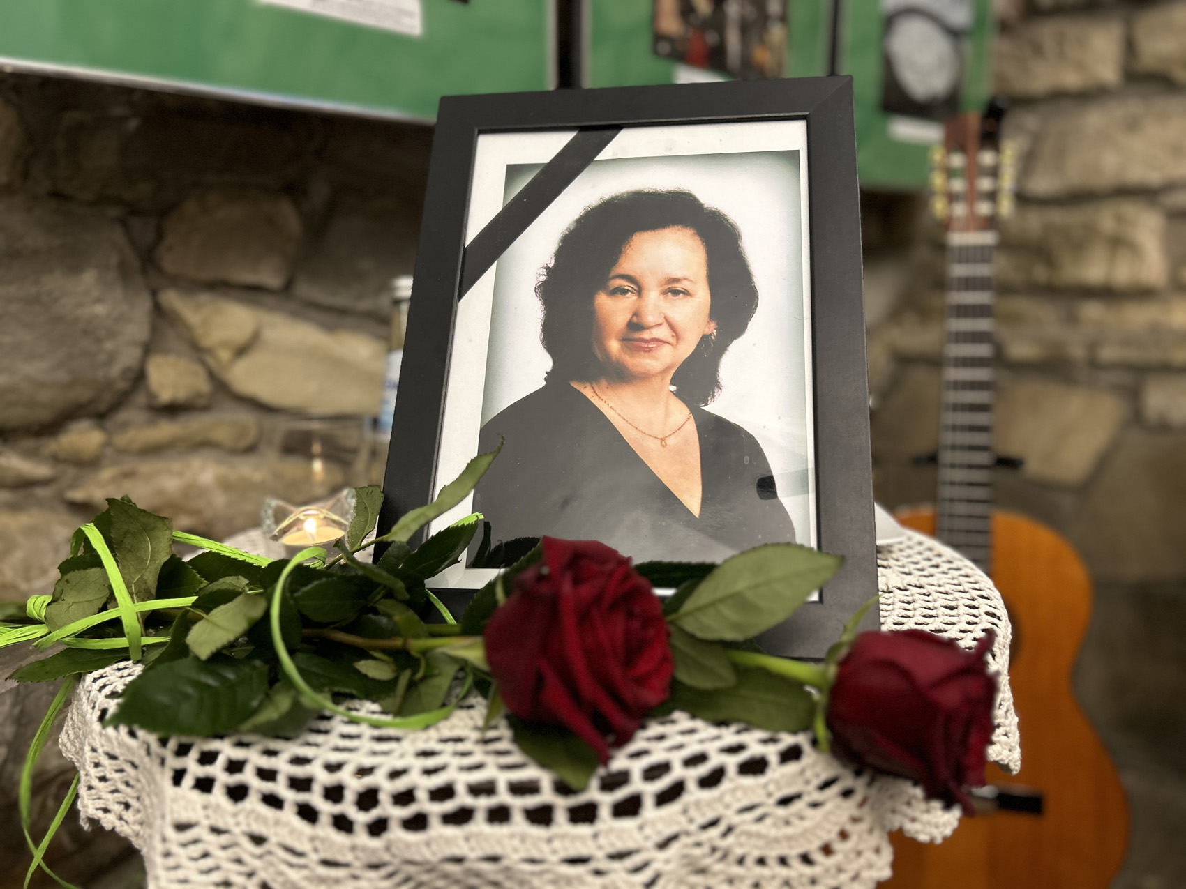 Wieczór wspomnień o Marii Piniańskiej (9) Zdjęcie Marii Piniańskiej w czarnej ramce na stoliku z serwetką. Przy ramce leż dwie czerwone róże oraz pali się mała świeczka. W tle widoczny jest kawałek ściany, zdjęcia oraz gitara.