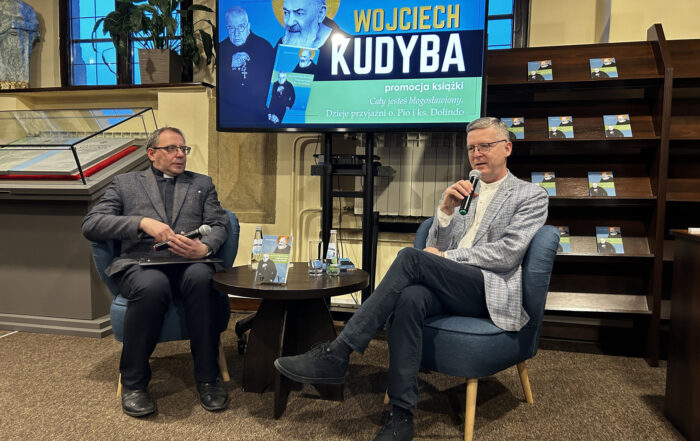 Dwóch mężczyzn siedzących przy stoliku: Wojciech Kudyba mówi do mikrofonu, prowadzący słucha. W tle regał z książkami Wojciecha Kudyby oraz ekran z wizerunkiem ojca Pio oraz księdza Dolindo.