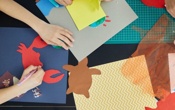 Zdjęcie przedstawia dłonie dzieci wykonywujące prace plastyczne. Jena dłoń trzyma w ręku nożyczki i wycina z żółtego papieru, inna dłoń trzyma długopis i pisze po wyciętym kształcie.
