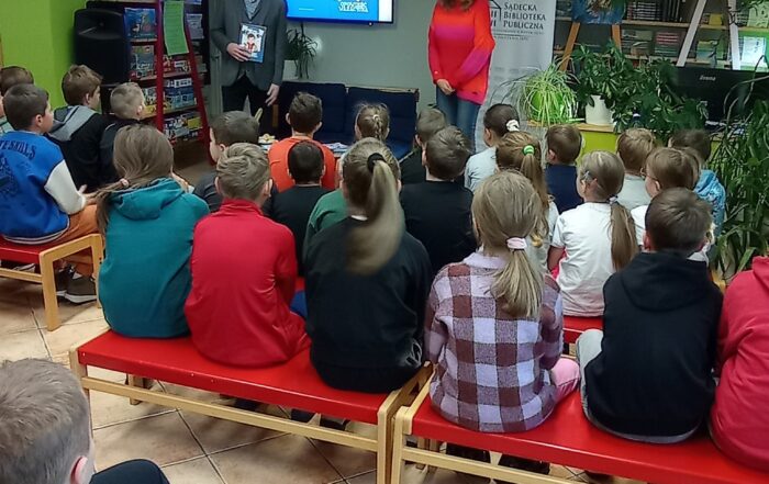 Uśmiechnięta kobieta w czerwono różowym swetrze oraz stojący obok niej mężczyzna w garniturze rozmawiają z dziećmi. Dookoła ustawione są regały z licznymi książkami. Dzieci z zainteresowaniem przysłuchują się.