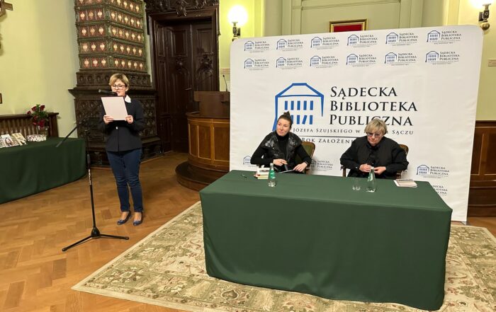 Sala ratusza. Po lewej stronie, przed mikrofonem stoi kobieta, trzymająca w ręku kartkę, z której czyta. Obok przy stoliku siedzą dwie kobiety, za nimi logo Sądeckiej Biblioteki Publicznej.
