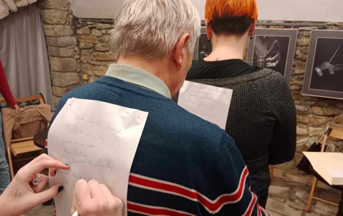 Dwie osoby z przypiętymi na plecach kartkami stoją jedna za drugą. Na pierwszym planie dłonie, które zapisują coś na kartce na plecach jednej z nich. W tle ściana z kamienia, na której wiszą biało-czarne fotografie.