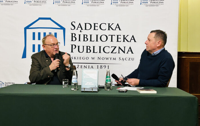 Dwóch mężczyzn siedzi za długim stołem. Stół nakryty jest zielonym obrusem. Na stole znajdują się: dwie szklanki, dwie butelki z wodą, książka stojąca na stojaku, dokumenty, leżąca książka. Mężczyzna pierwszy z lewej trzyma w ręce mikrofon blisko ust, mówi do niego. Patrzy na mężczyznę siedzącego obok. Drugi mężczyzna patrzy na mówiącego, słucha. W jednej ręce trzyma mikrofon, w drugiej okulary. W tle za siedzącymi duża biała ścianka z logotypem Sądeckiej Biblioteki Publicznej.