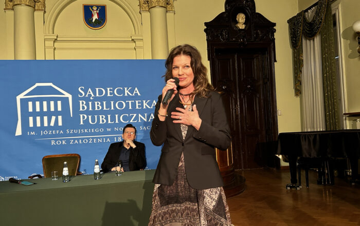Elegancka sala ratuszowa. Kobieta w sukience stoi z mikrofonem w dłoni. Za nią siedzi mężczyzna przy zielonym stole. W tle widać ogromne, drewniane drzwi oraz fortepian a także logo Sądeckiej Biblioteki Publicznej.