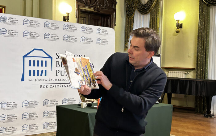 Na zdjęciu widoczny jest mężczyzna, trzymający w ręku książkę. Po lewej stronie widnieje logo Sądeckiej Biblioteki Publicznej.