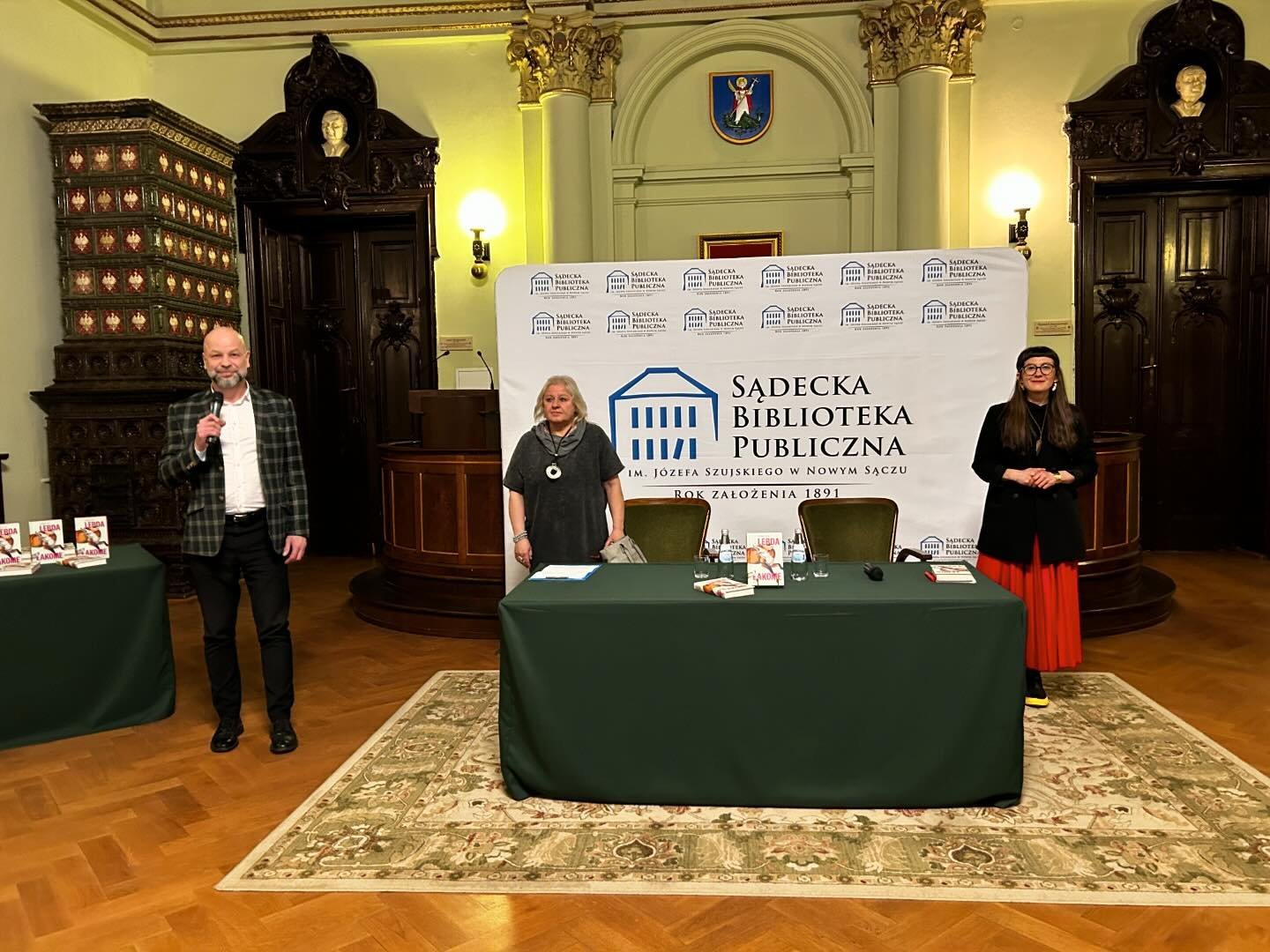 Małgorzata Lebda_1 W eleganckiej sali ratusza po lewej stronie stoi mężczyzna z mikrofonem w ręku, po prawej stronie stoją dwie kobiety przy stole z krzesłami. za nimi widnieje duże logo Sądeckiej Biblioteki Publicznej.