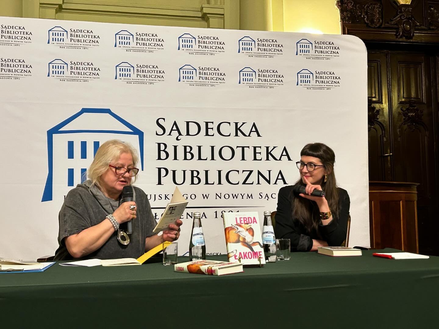 Małgorzata Lebda_2 Dwie kobiety siedzą przy stole. Kobieta po prawej stronie czyta książkę do mikrofonu. Kobieta po prawej stronie przysłuchuje się. Przed nimi leżą książki oraz napoje.