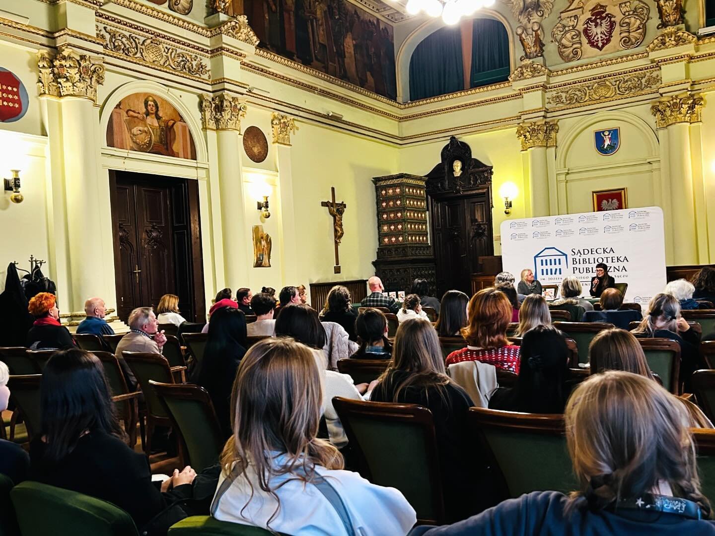 Małgorzata Lebda_3 Elegancka sala ratusza. Ludzie siedzący na krzesłach słuchają kobiety siedzącej przed nimi przy stole. Obok niej po prawej stronie przy stole siedzi druga kobieta.