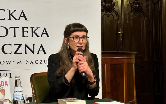 Kobieta siedzi przy stole, w dłoniach trzyma mikrofon do którego mówi. Przed nią leży książka oraz woda mineralna.