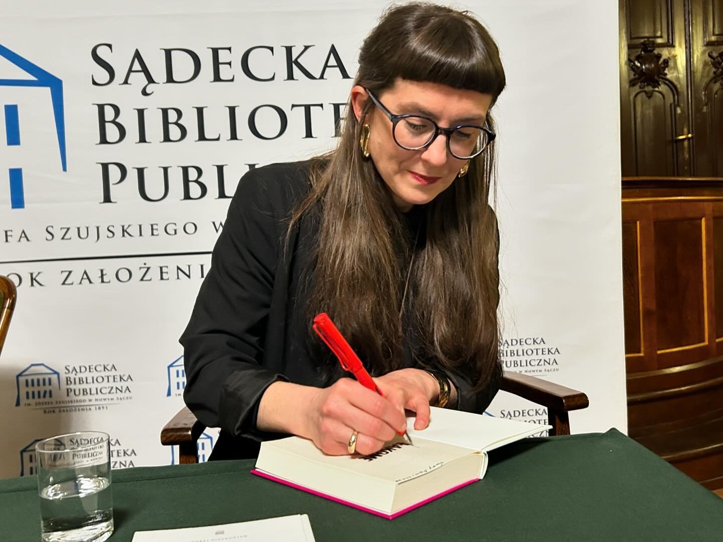 Małgorzata Lebda_5 Kobieta w długich, ciemnych włosach, ubrana na czarno, w okularach siedzi przy stole i podpisuje książkę czerwonym długopisem.