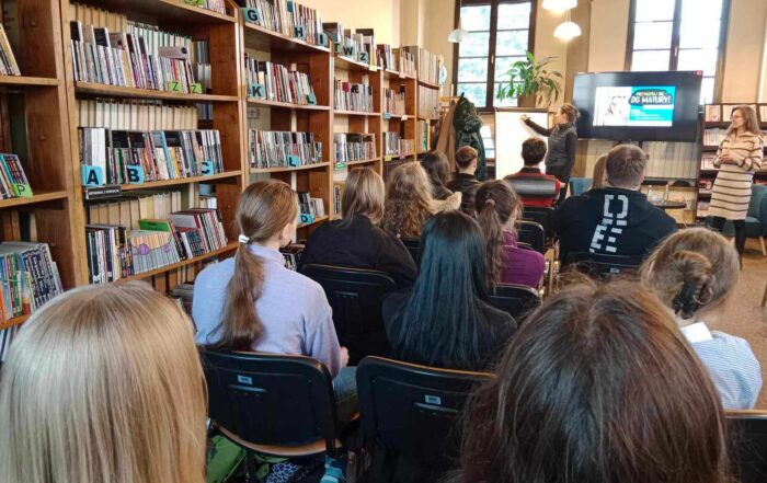 Uczniowie szkół siedzą na krzesłach, słuchają kobiety stojącej przy tablicy. Po bokach, przy ścianach ustawione są regały z książkami.